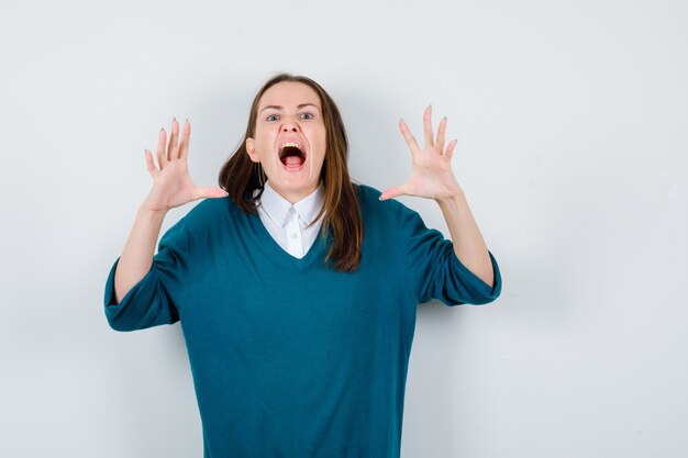 Young woman showing claws imitating a cat in sweater over white shirt and looking aggressive. front view.