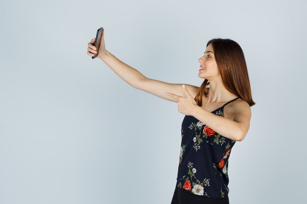 Young woman showing gun gesture while taking selfie on smartphone in blouse and looking confident , front view.