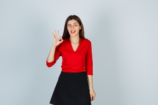 Young woman showing ok sign, sticking tongue out in red blouse