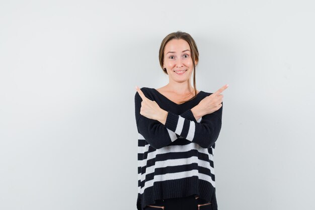 Young woman showing opposite directions with index fingers in striped knitwear and black pants and looking happy