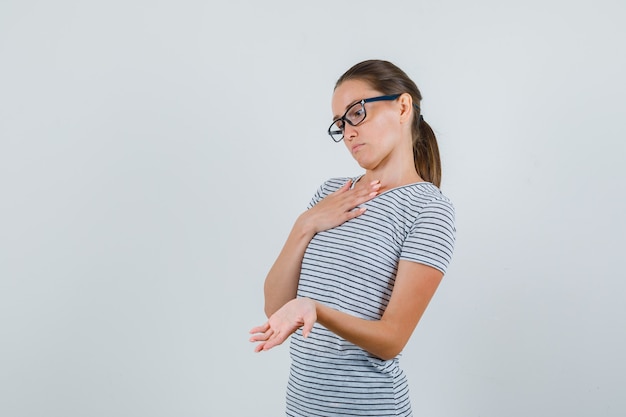 Young woman showing palm in striped t-shirt, glasses and looking pensive. front view.