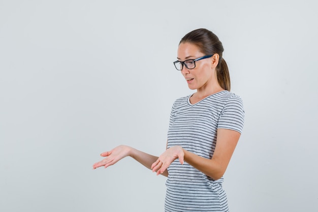 Young woman showing palms in striped t-shirt, glasses and looking amazed. front view.