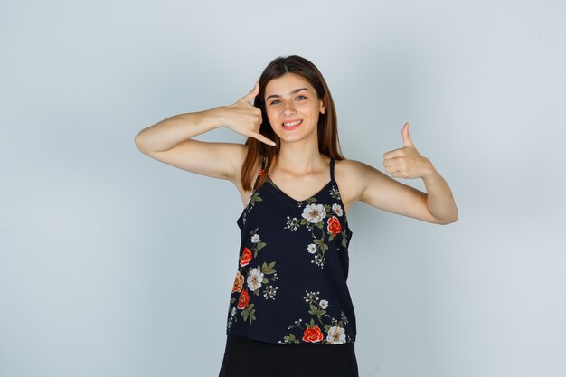 Young woman showing phone gesture and thumb up in blouse, skirt and looking merry. front view.