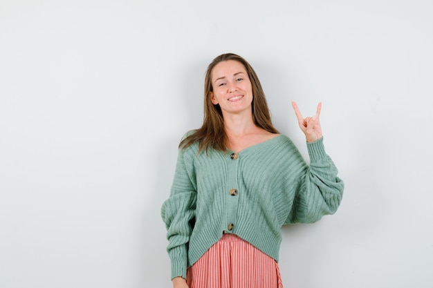 Young woman showing rock gesture in cardigan, skirt and looking happy. front view.