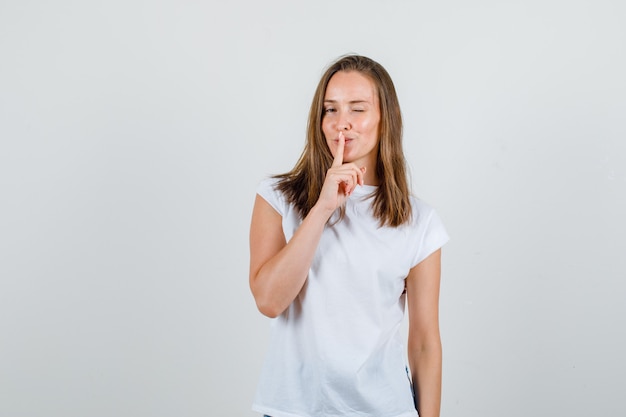 Young woman showing silence gesture with blinked eye in white t-shirt front view.