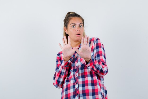 Young woman showing stop gesture in casual shirt and looking scared , front view.