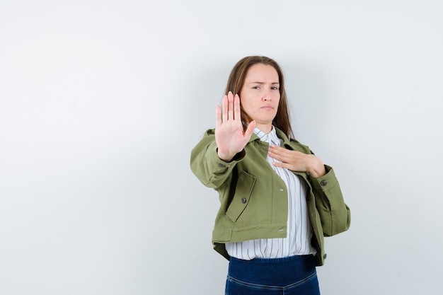 Young woman showing stop gesture in shirt, jacket and looking resolute , front view.