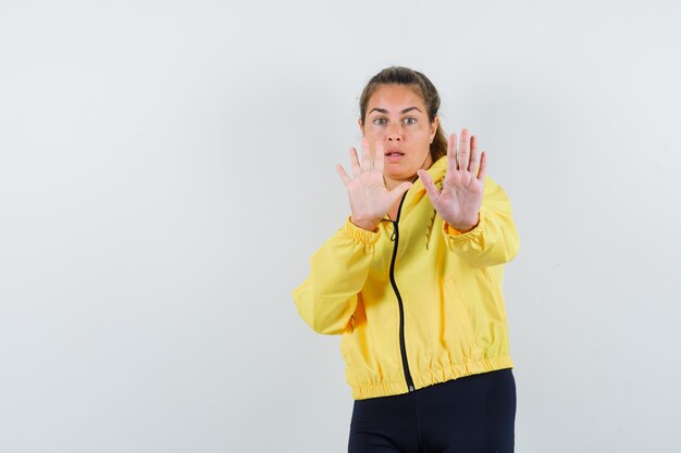 Young woman showing stop sign with both hands in yellow bomber jacket and black pants and looking scared