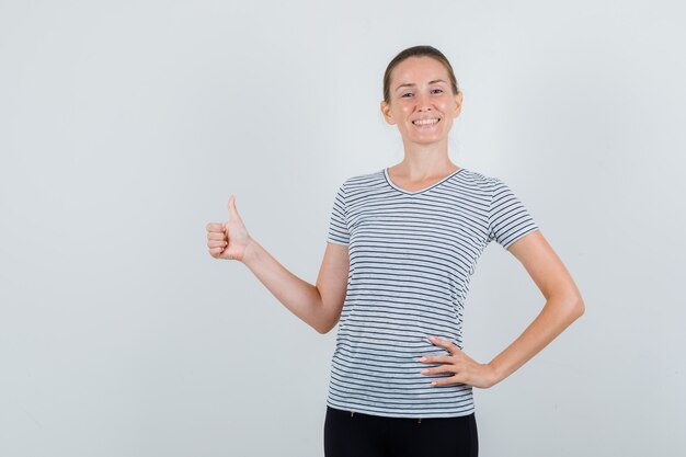 Young woman showing thumb up in t-shirt, pants and looking cheerful , front view.