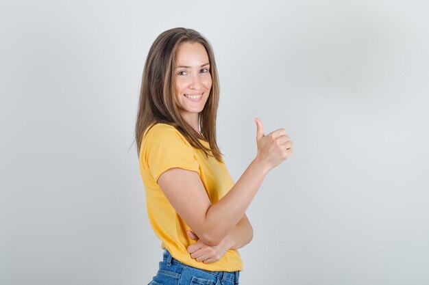 Young woman showing thumb up in yellow t-shirt, shorts and looking cheerful