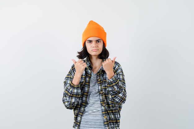 Young woman showing thumbs up in orange hat checkered shirt looking serious 