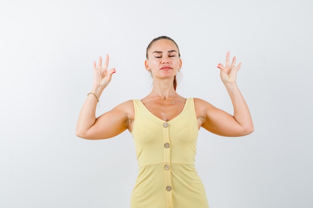 Free photo young woman showing yoga gesture with shut eyes in yellow dress and looking relaxed. front view.