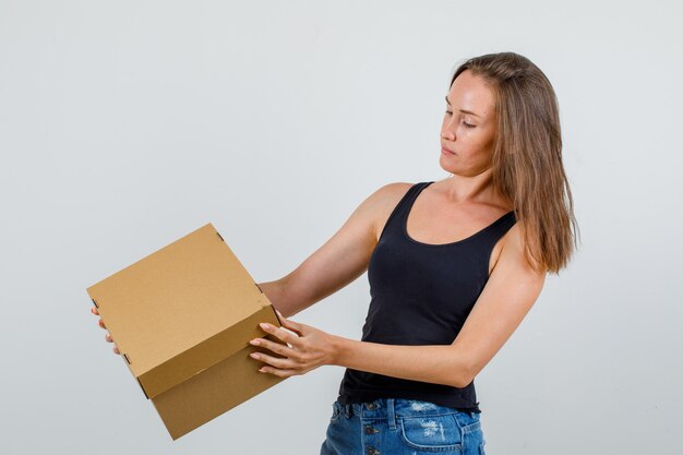 Young woman in singlet, shorts holding cardboard box and looking careful