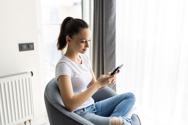 Free photo young woman sit in chair use smartphone wear casual style clothes in house indoors