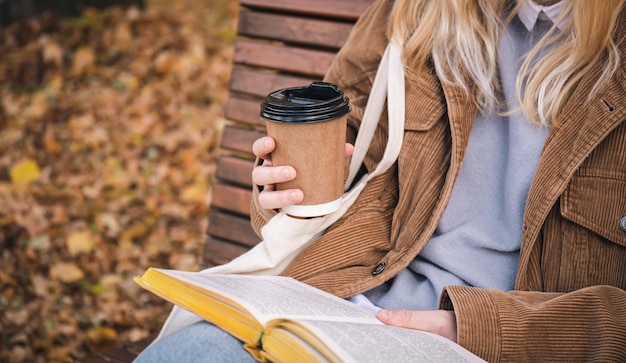 Free Photo a young woman sits on a bench in an autumn park drinks coffee and reads a book