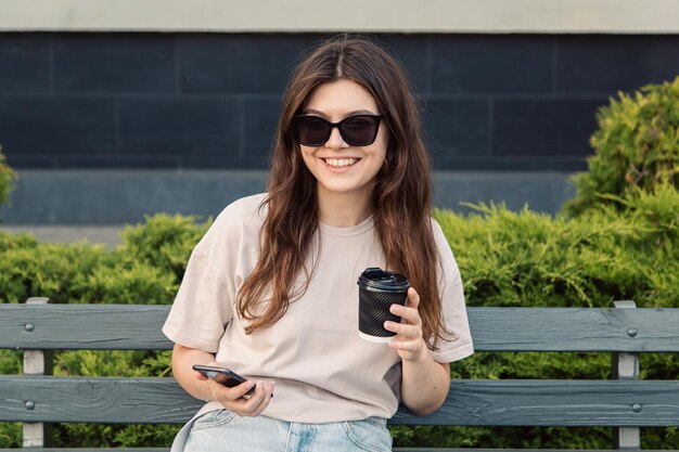 A young woman sits on a bench with a cup of coffee and a smartphone