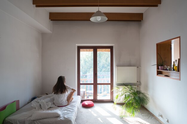 Young woman sitting on bed and looking through the window.