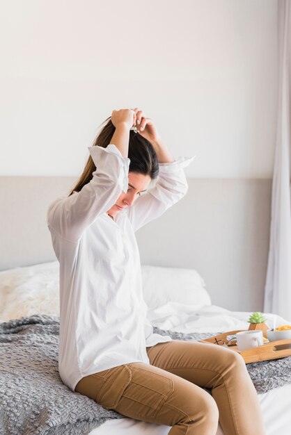 Young woman sitting on bed tying her hair