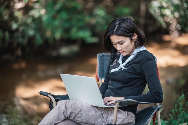 Young woman sitting on camping chair and use laptop computer while relax on camping in forest copy space