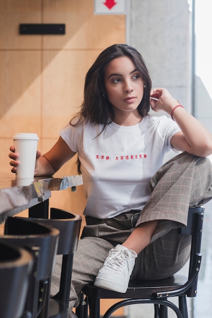 Free photo young woman sitting in chair holding disposable coffee cup at table