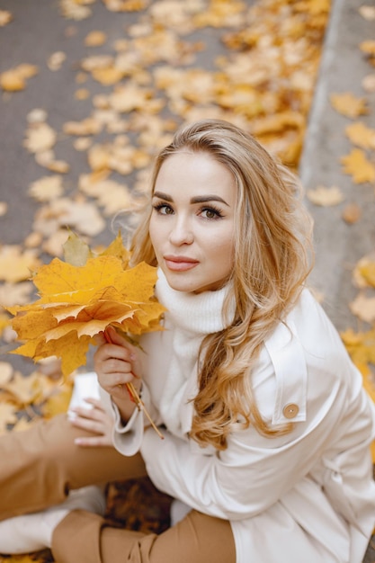 Young woman sitting on a curb in autumn forest. Blonde woman holding a yellow leaves. Girl wearing beige coat and brown trousers.