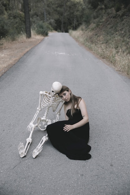 Young woman sitting on empty road in forest with skeleton