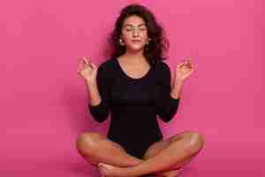 Free photo young woman sitting in lotus position with closed eyes, doing yoga, relaxing at home, wearing black combidress, having wavy hair, sitting on floor on pink concentrated wall.