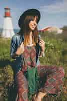 Free photo young woman sitting in nature, lighthouse, bohemian outfit, denim jacket, black hat, smiling, happy, summer, stylish accessories