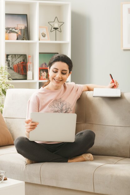 Young woman sitting on sofa behind coffee table holding and used laptop writes on book in living room