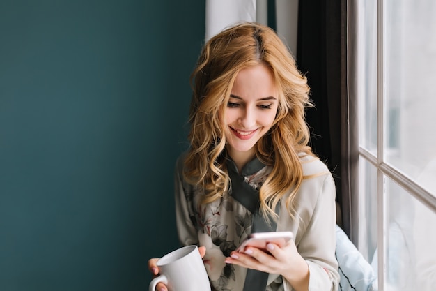 Free Photo young woman sitting next to window and texting on phone while drinking morning coffee or tea. her blonde hair is wavy. she is in bright room with turquoise wall. wearing silk pajama.