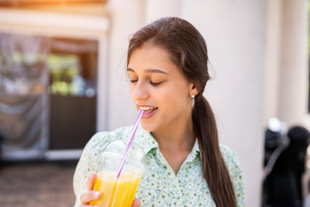 Free photo young woman smiling and drinking cocktail with ice in plastic cup with straw on city street.