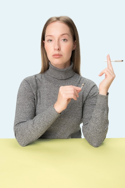 The young woman smoking cigarette while sitting at table at studio. Trendy colors. The portrait of caucasian girl in minimalism style with copy space