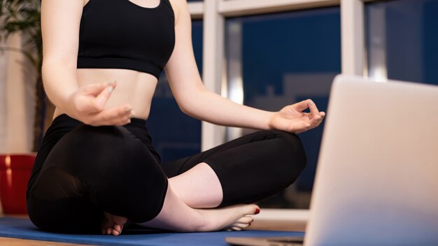 Young woman in sportswear is meditating on a yoga mat with laptop in front of her