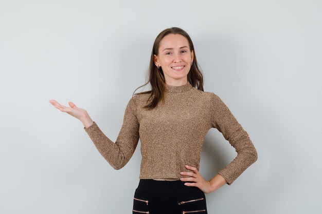 Young woman spreading palm aside in golden blouse and looking merry , front view.