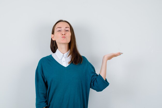 Young woman spreading palm aside, pouting lips in sweater over white shirt and looking peaceful , front view.