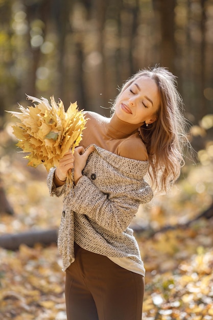 Young woman standing in autumn forest. Brunette woman holding a yellow leaves. Girl wearing fashion brown jacket.