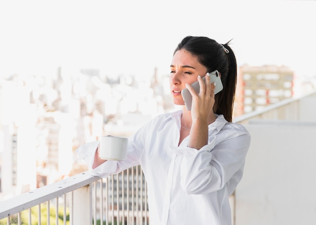 Young woman standing in balcony holding cup of coffee talking on mobile phone