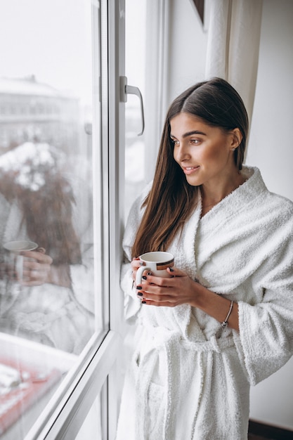 Young woman standing by the window drinking hot coffee