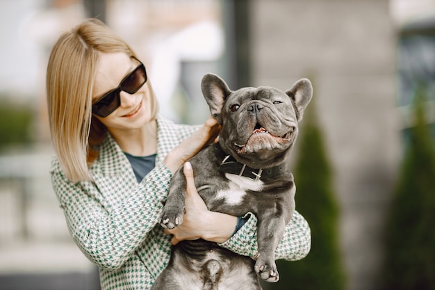 Free photo young woman standing near cafe outdoors holding black french bulldog. girl wearing black sunglasses, shorts and grey jacket