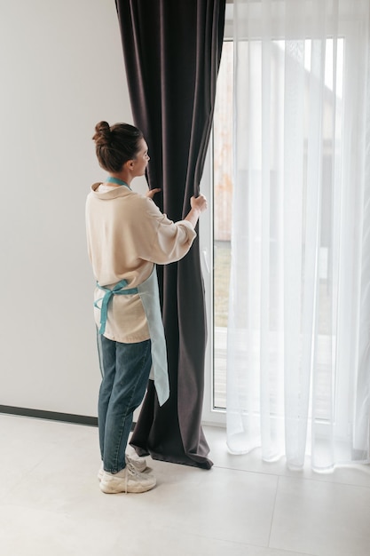 Free photo young woman standing near the widnow and fixing the curtains