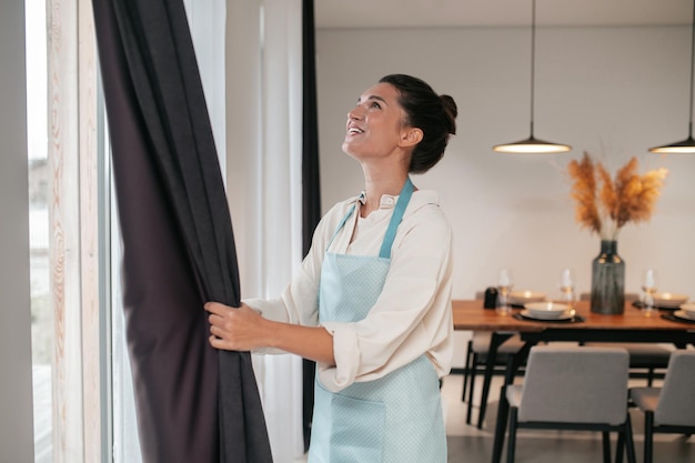 Free photo young woman standing near the widnow and fixing the curtains