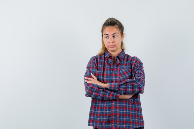 Young woman standing straight, looking away and posing at front in checked shirt and looking pensive