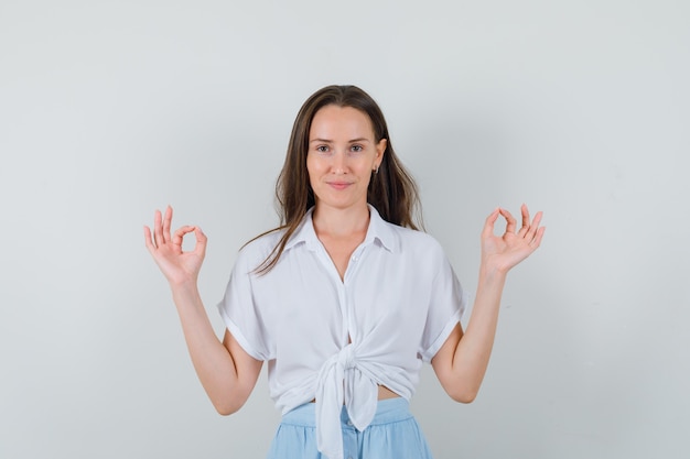 Young woman standing straight, showing ok sign with both hands in white blouse and light blue skirt and looking confident