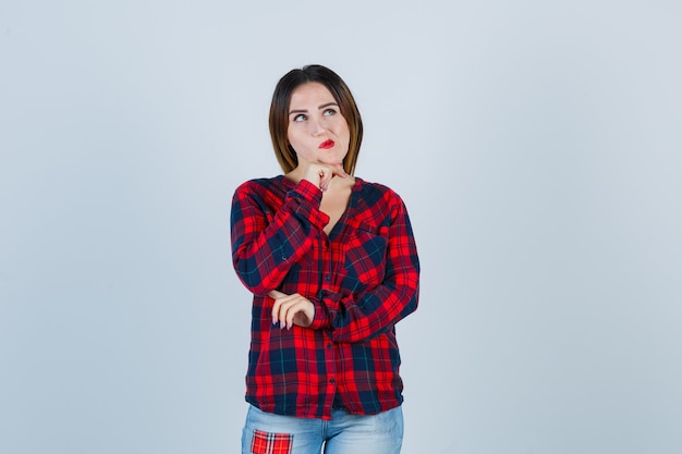 Young woman standing in thinking pose, propping chin on hand in checked shirt, jeans and looking pensive. front view.