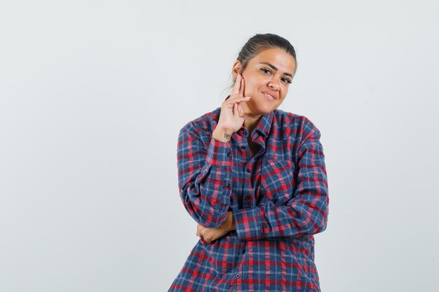 Young woman standing in thinking pose while holding hand on cheek in checked shirt and looking happy. front view.