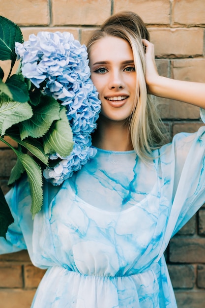 Young woman in straw hat holding blue hydrangea flowers standing agaist breek street wall