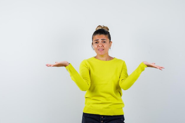 Young woman stretching hands in questioning manner in yellow sweater and black pants and looking puzzled 