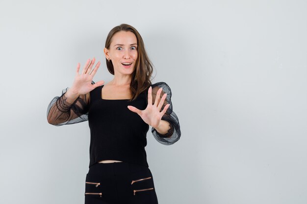 Young woman stretching hands to stop something in black blouse and black pants and looking happy. front view.