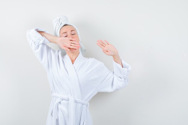 Young woman stretching upper body while yawning in white bathrobe, towel and looking relaxed , front view.