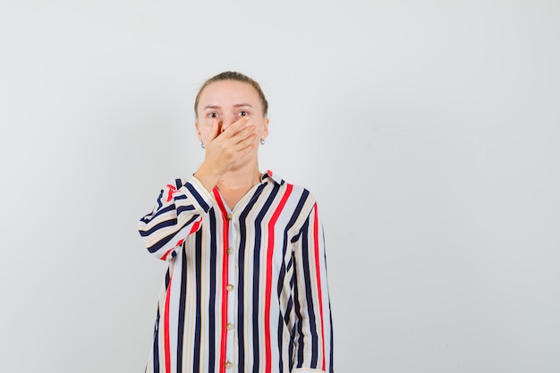 Young woman in striped blouse covering her mouth with both hands and looking surprised